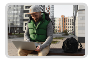 man in sporty outfit sitting on a outside bench in middle of a big city, using laptop computer